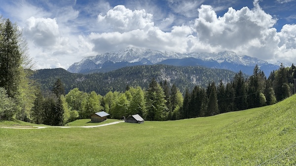 Widok na polanę, drewniane chatki, las i Alpy w chmurach